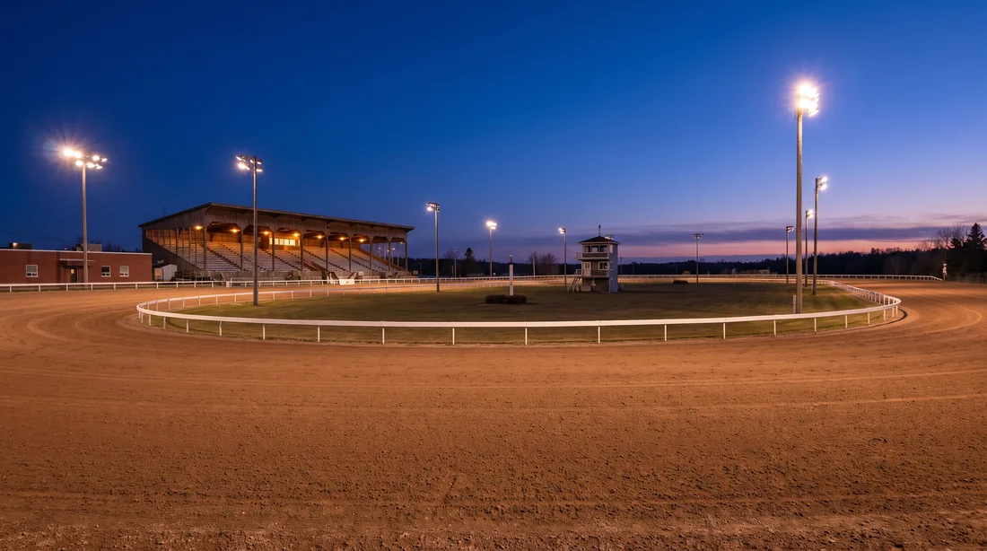 An empty harness racing oval at blue hour: reddish dirt, a white rail, a lit covered grandstand, light towers and a judges tower in the centre field.